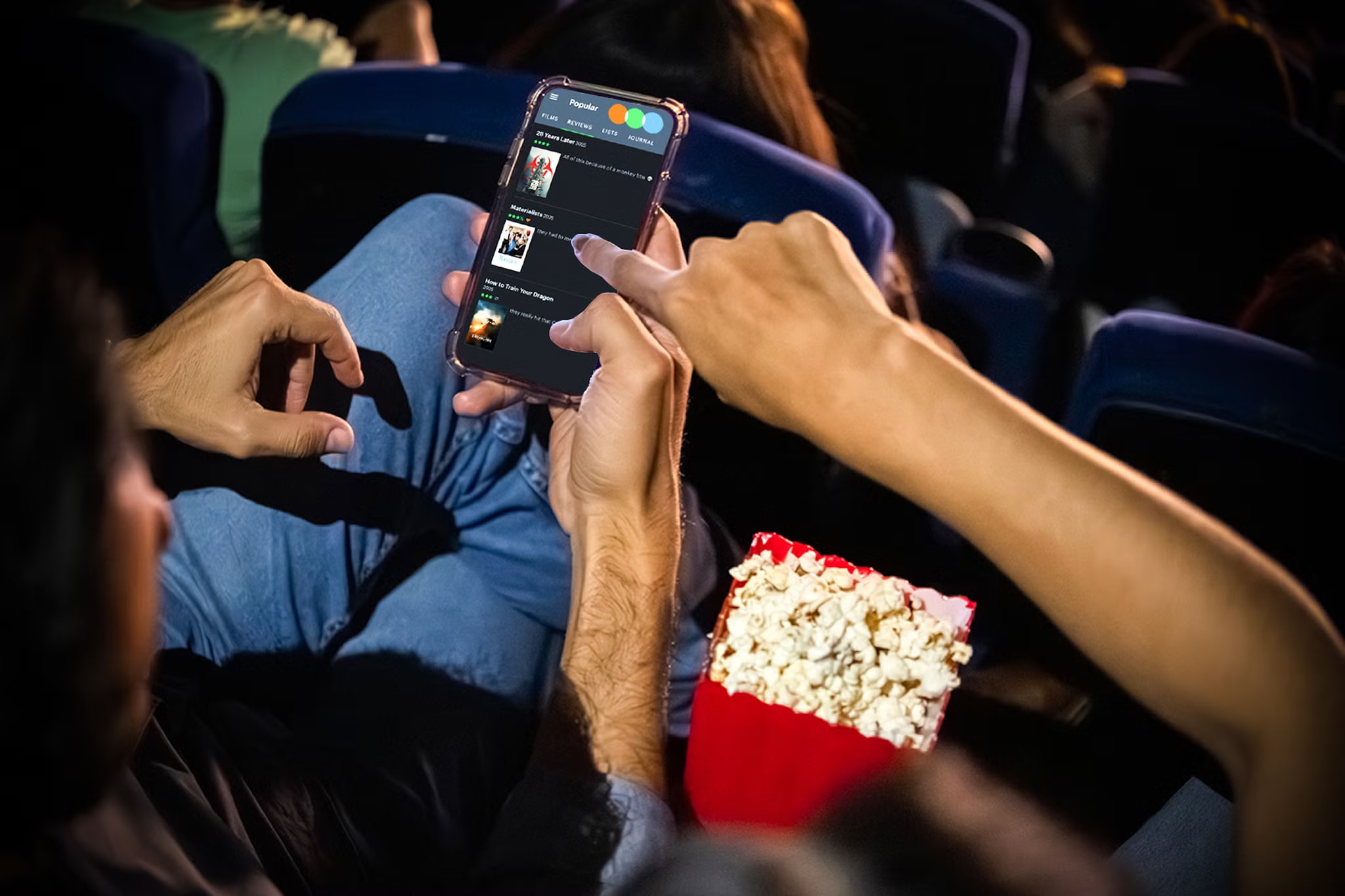 Moviegoers sit in a theater while one person scrolls through Letterboxd on a smartphone beside a box of popcorn.