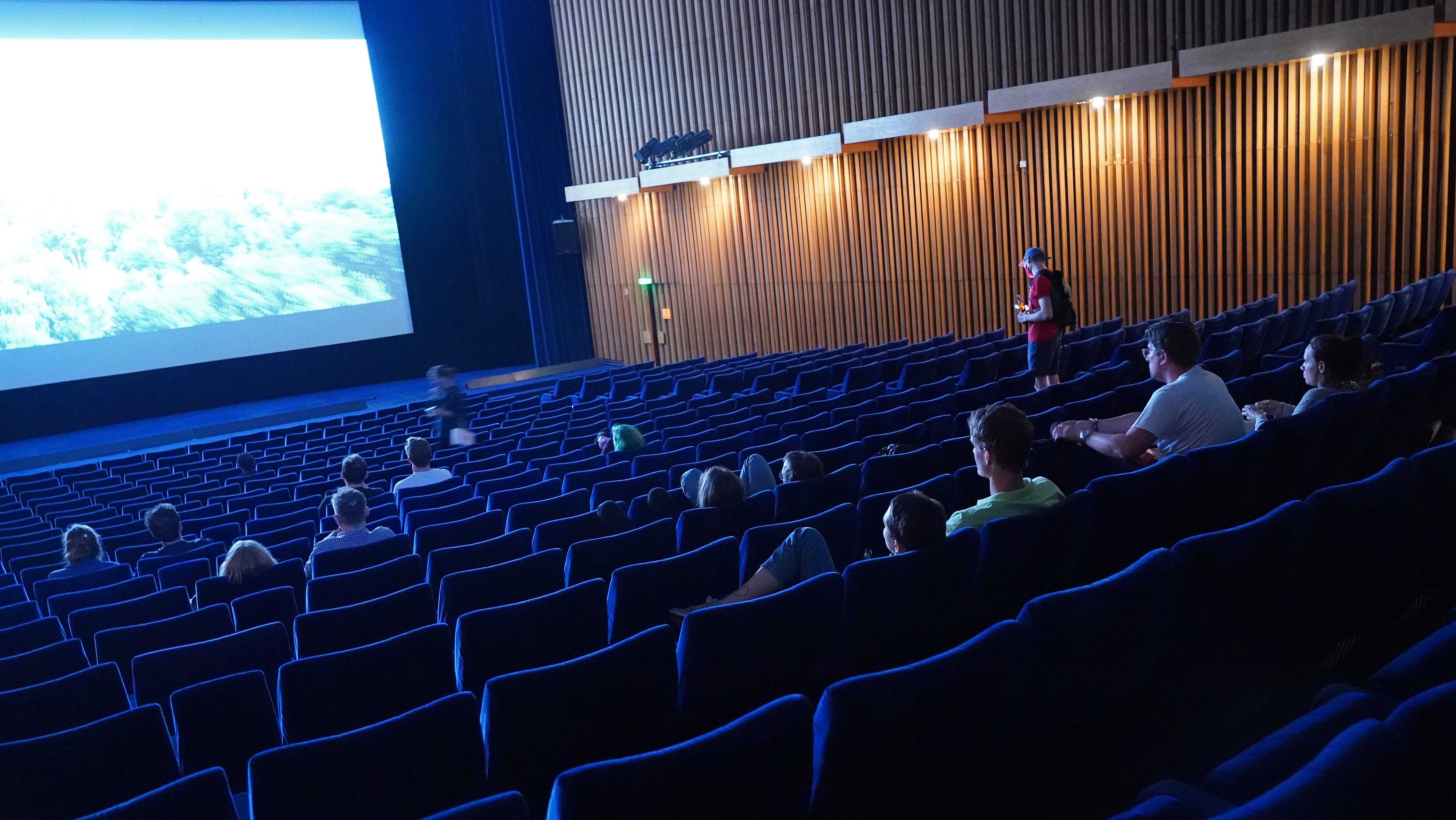 A modern cinema auditorium with many empty blue seats and a small audience waiting for a screening.