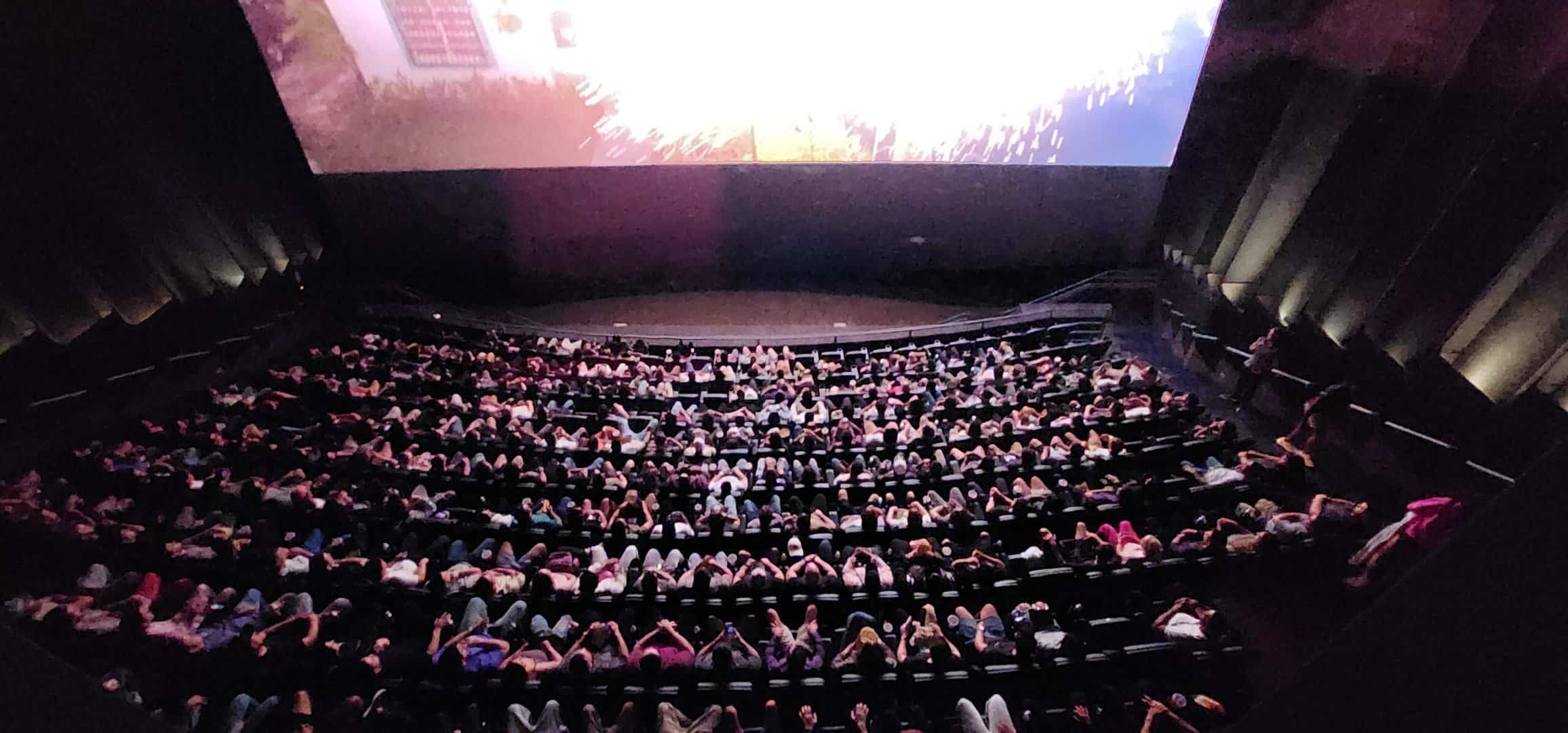 A packed movie theater viewed from the front beneath a brightly illuminated screen.