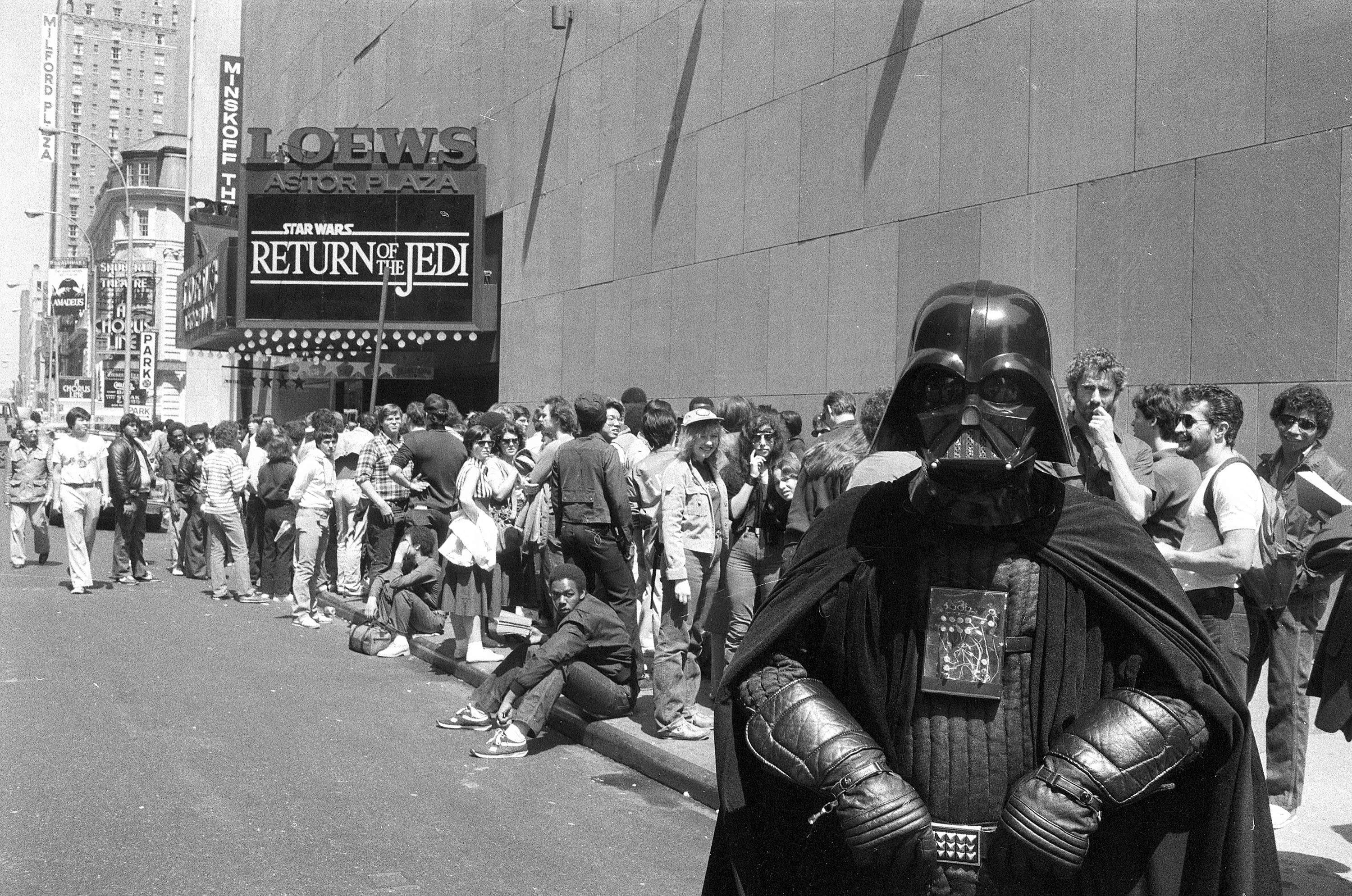 A large crowd waits outside a theater showing Return of the Jedi as a person dressed as Darth Vader stands in the foreground.