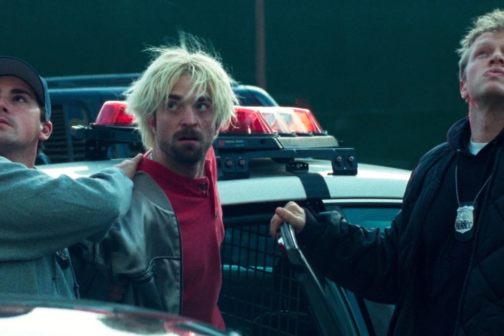 Robert Pattinson, with bleached hair and a beard, stands beside a police car in Good Time.
