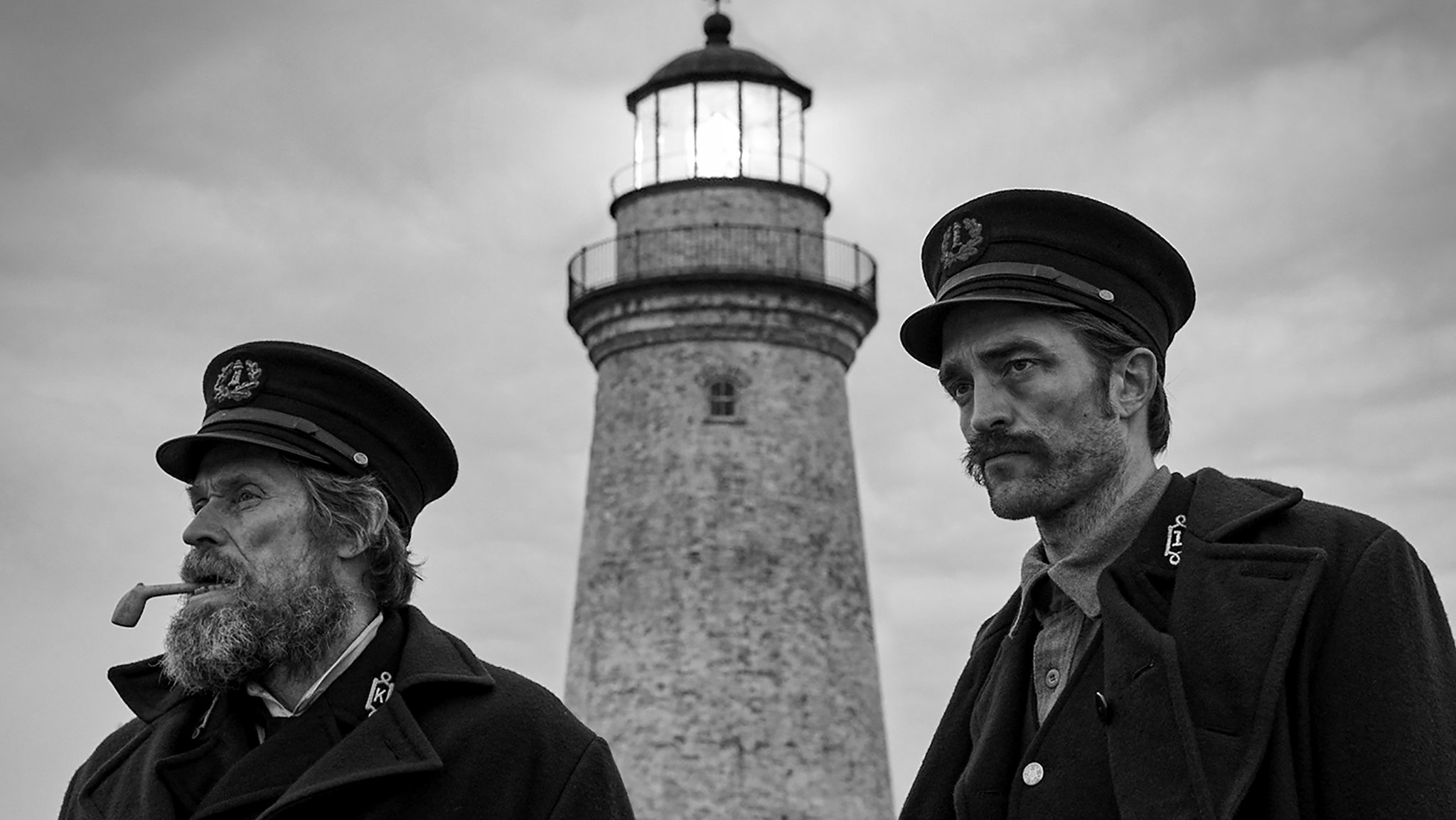 Willem Dafoe and Robert Pattinson stand in period lighthouse-keeper uniforms beneath a lighthouse.