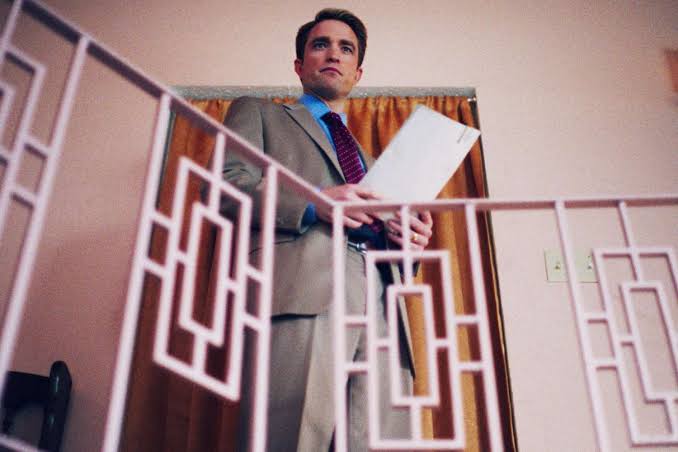 Robert Pattinson stands on an interior staircase in a suit while holding papers in Primetime.