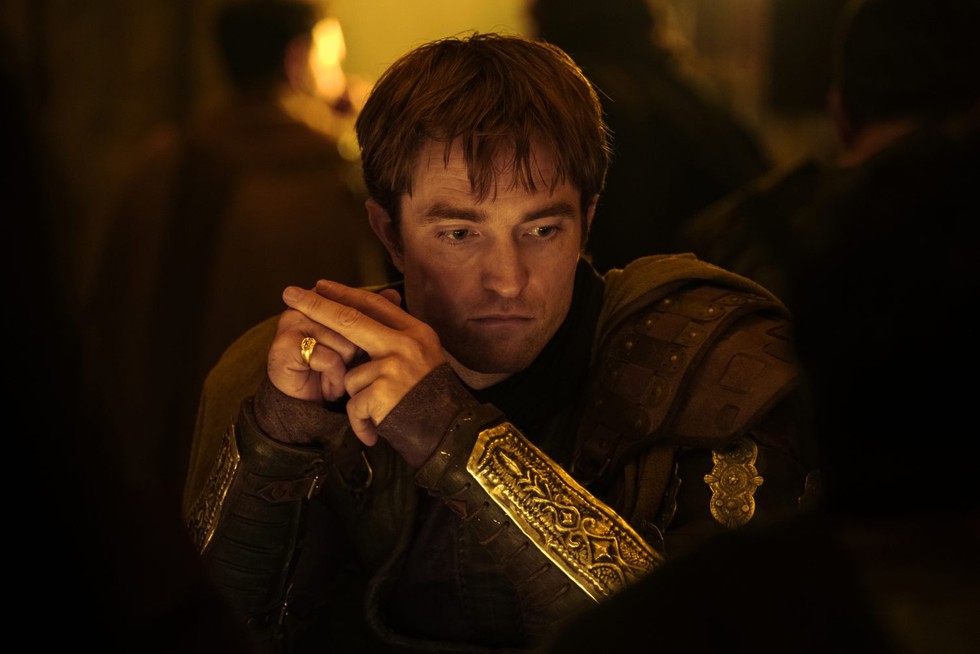 Robert Pattinson as Antinous sits in bronze armor with his hands folded.