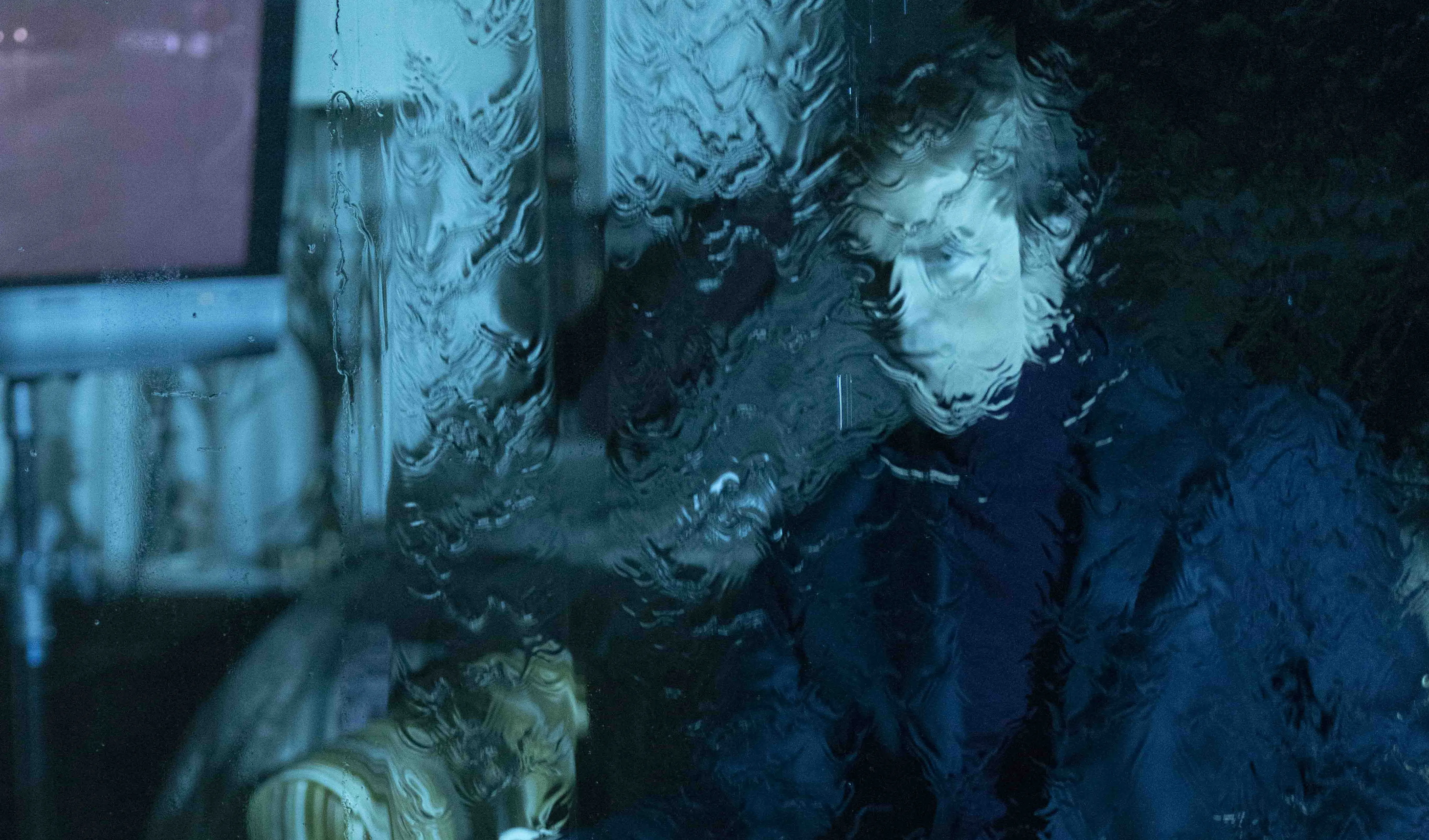 Robert Pattinson appears distorted behind rain-covered or textured glass in a blue-toned image from Primetime.
