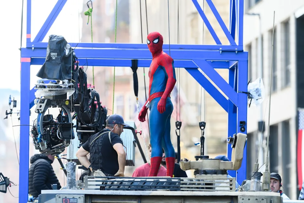 Spider-Man stands on a raised practical set beside a large camera rig during production.