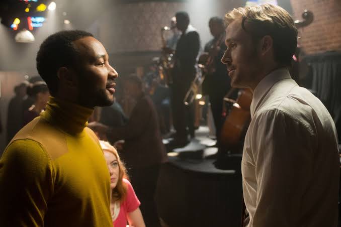 Sebastian speaks with a musician inside a crowded jazz club while Mia sits nearby.