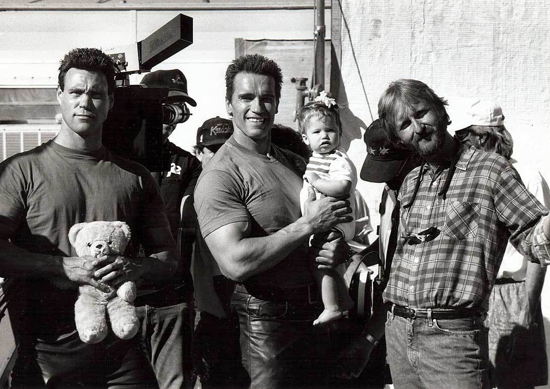 Arnold Schwarzenegger holds a child on the set of Terminator 2 while James Cameron stands nearby.