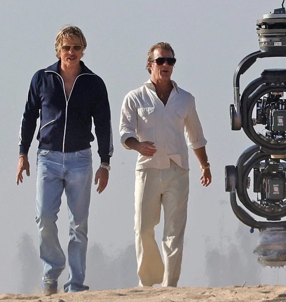 Brad Pitt walks across an outdoor film set beside another performer and a large camera rig during production of The Further Mis-Adventures of Cliff Booth.