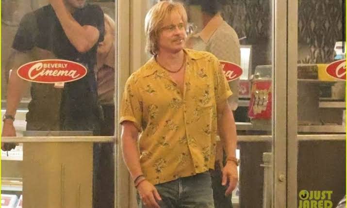 Brad Pitt as Cliff Booth stands outside a theater dressed in a yellow patterned shirt during production of The Further Mis-Adventures of Cliff Booth.