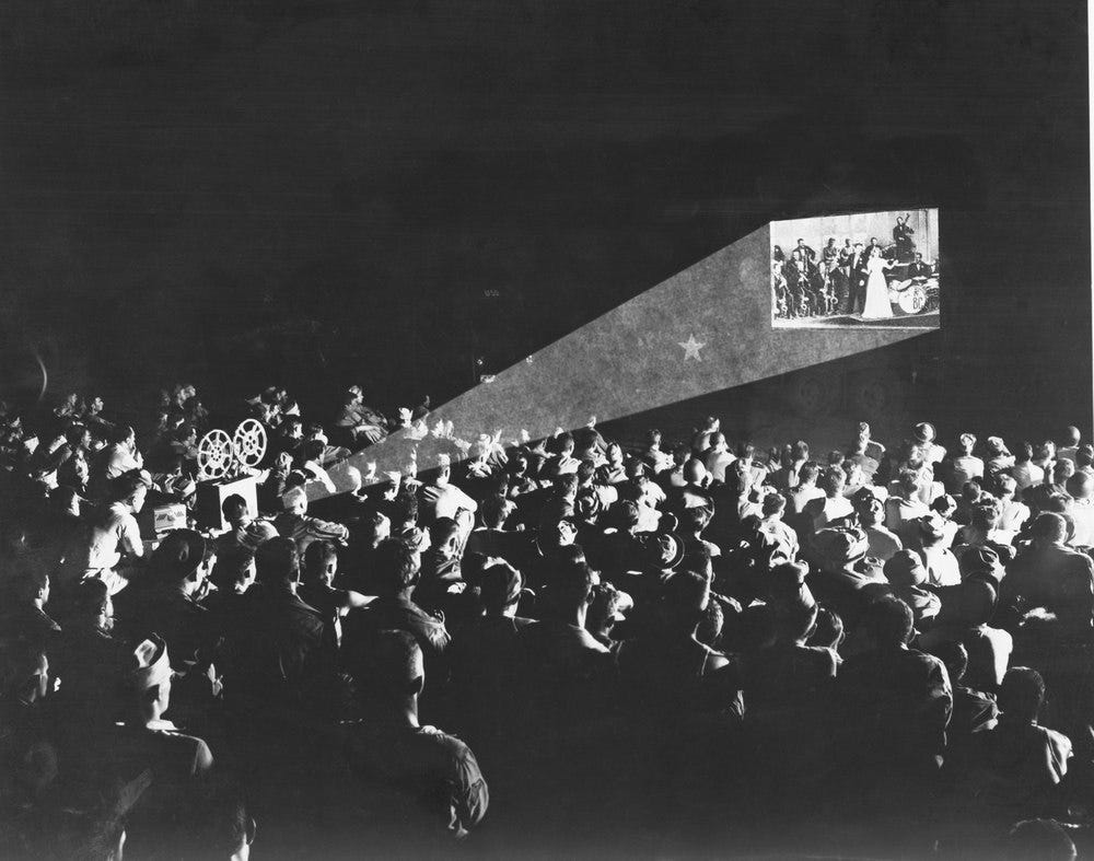 A black-and-white historical photograph shows a large audience watching a projected film in a theater.