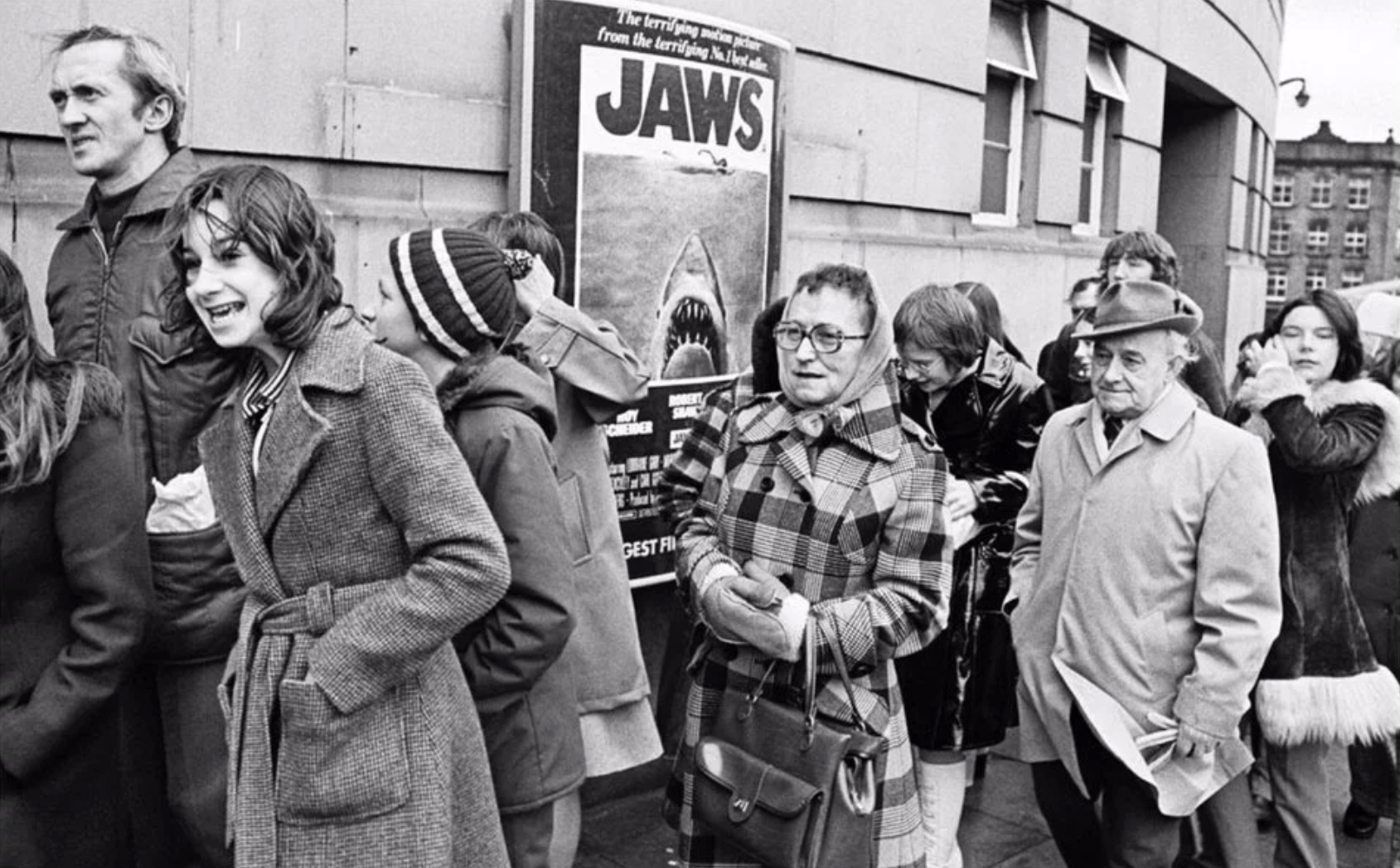 Black-and-white photograph of moviegoers waiting outside a theater with a Jaws poster visible behind them.