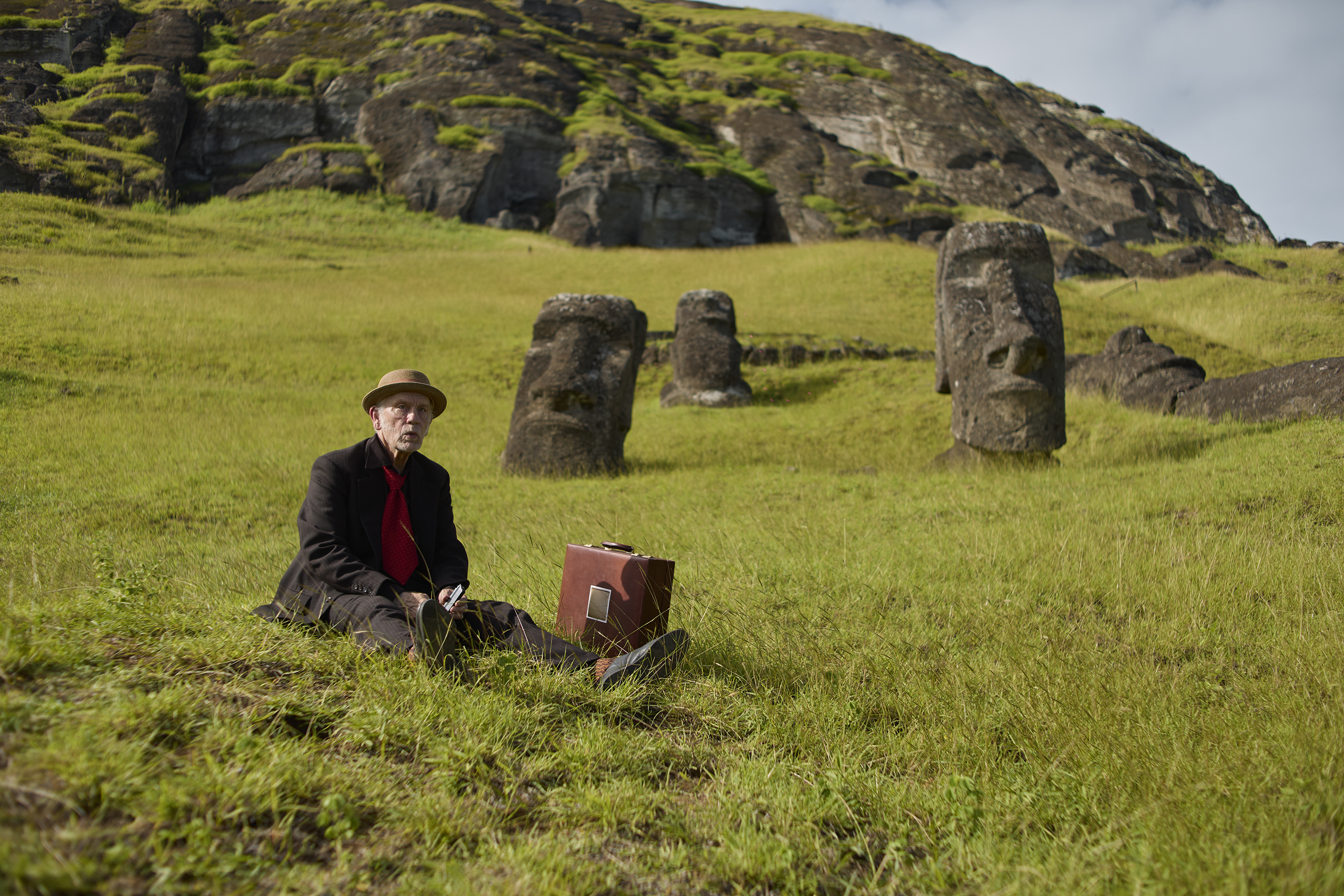 John Malkovich sits in the grass among the moai statues of Rapa Nui.