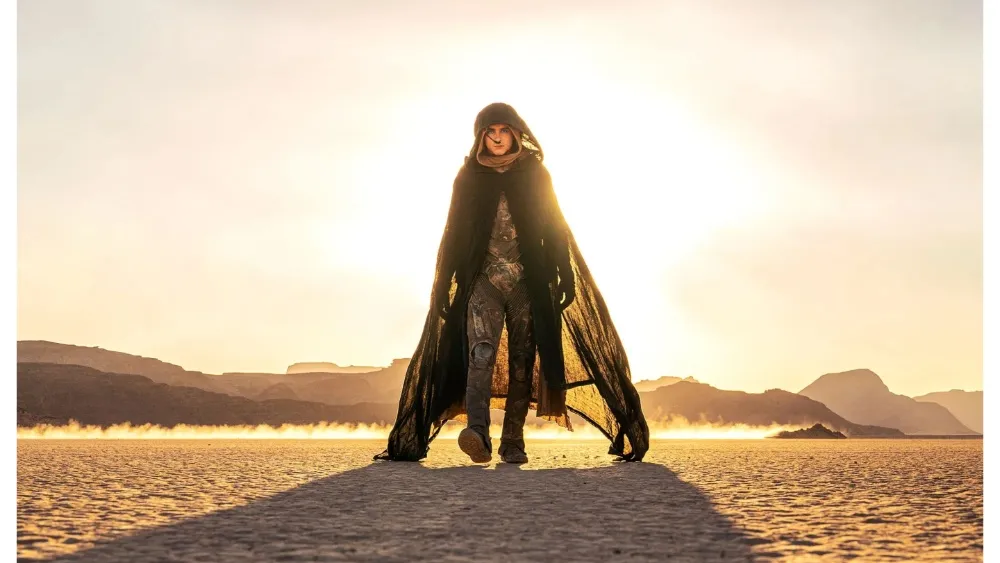 A cloaked figure walks across a desert landscape with bright sunlight behind them.