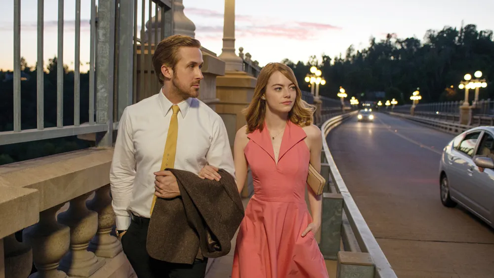 Ryan Gosling and Emma Stone walk together across a bridge at dusk in La La Land.