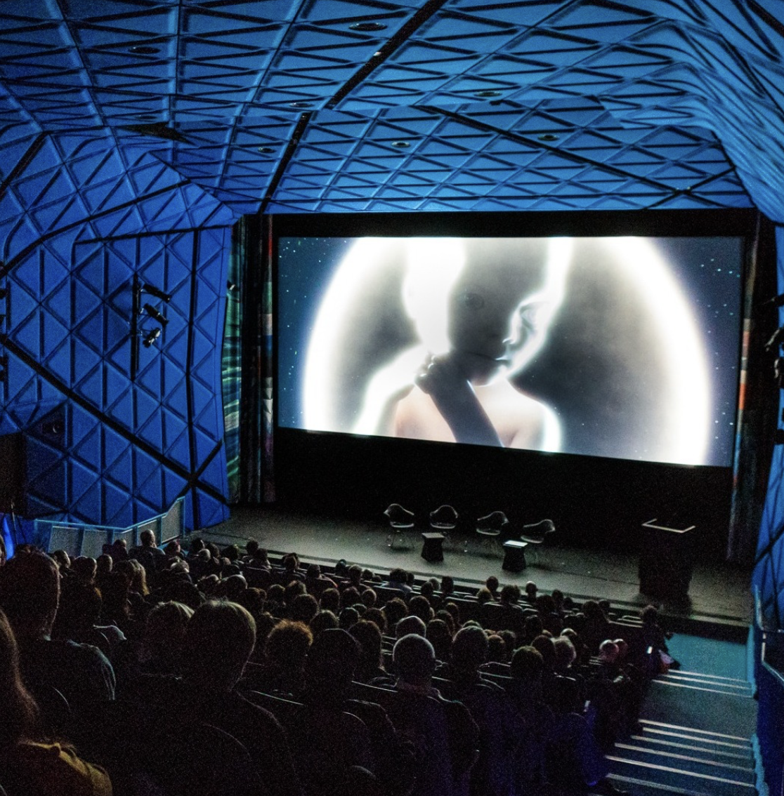 A large audience sits in a distinctive blue-lit cinema facing a bright movie screen.