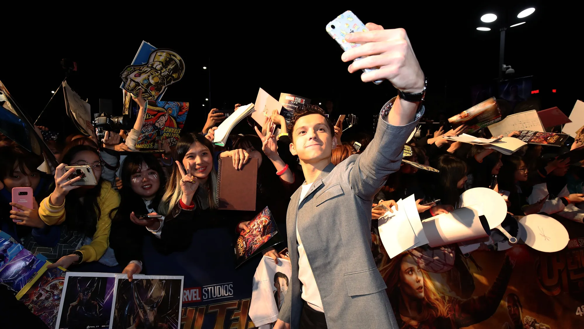 A film star takes a selfie with a dense crowd of fans holding posters and phones.