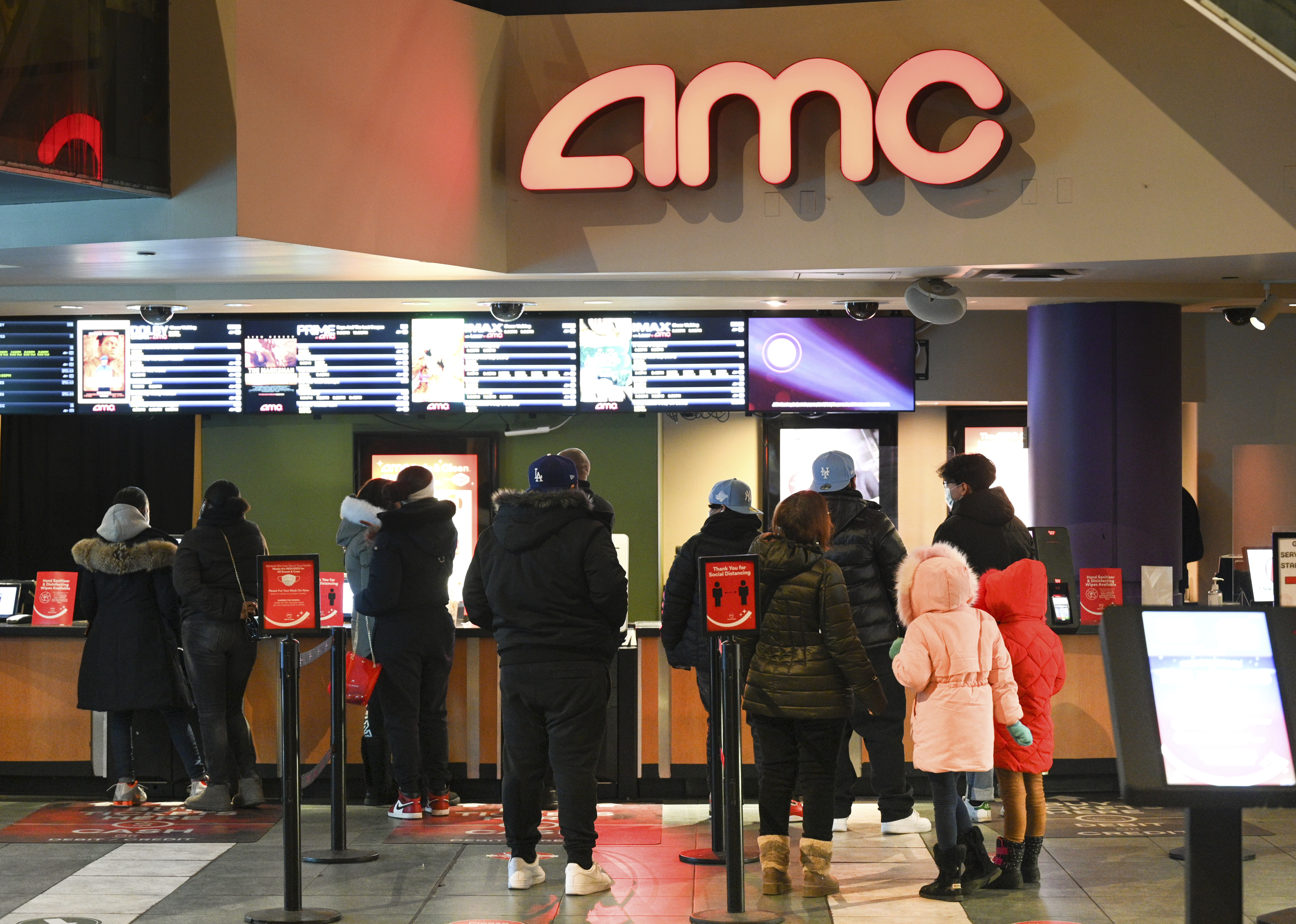 Moviegoers stand in line beneath digital showtime boards inside an AMC theater.