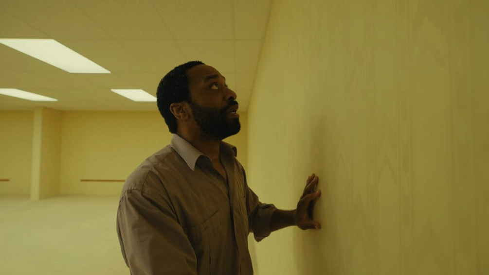 A man presses his hand against the wall of an empty yellow corridor.