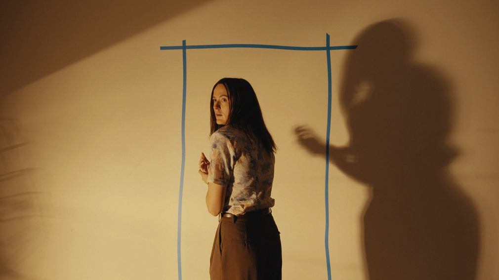A woman stands beside a taped rectangle on a bare yellow wall as a shadow looms behind her.