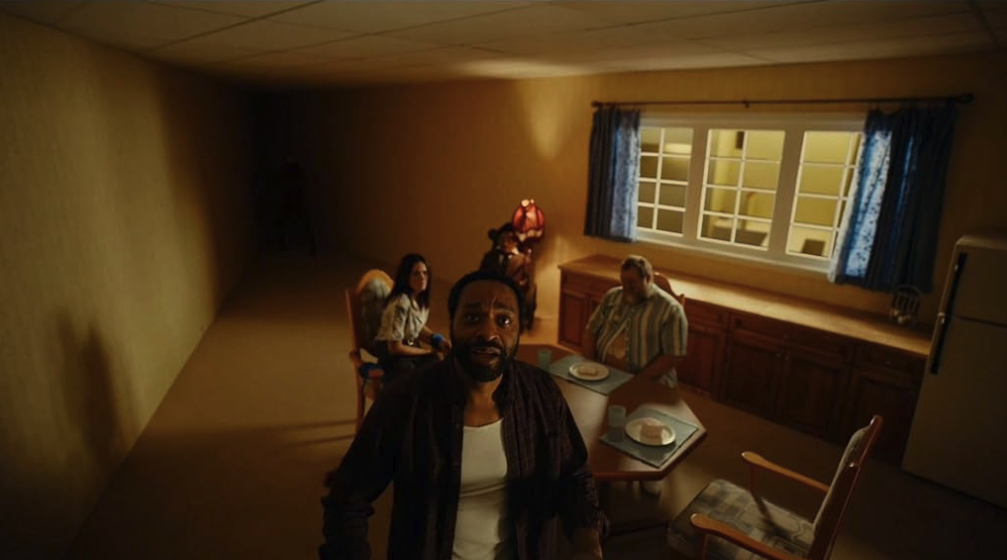 Figures gather in a dim, sparsely furnished room lit by a single window.