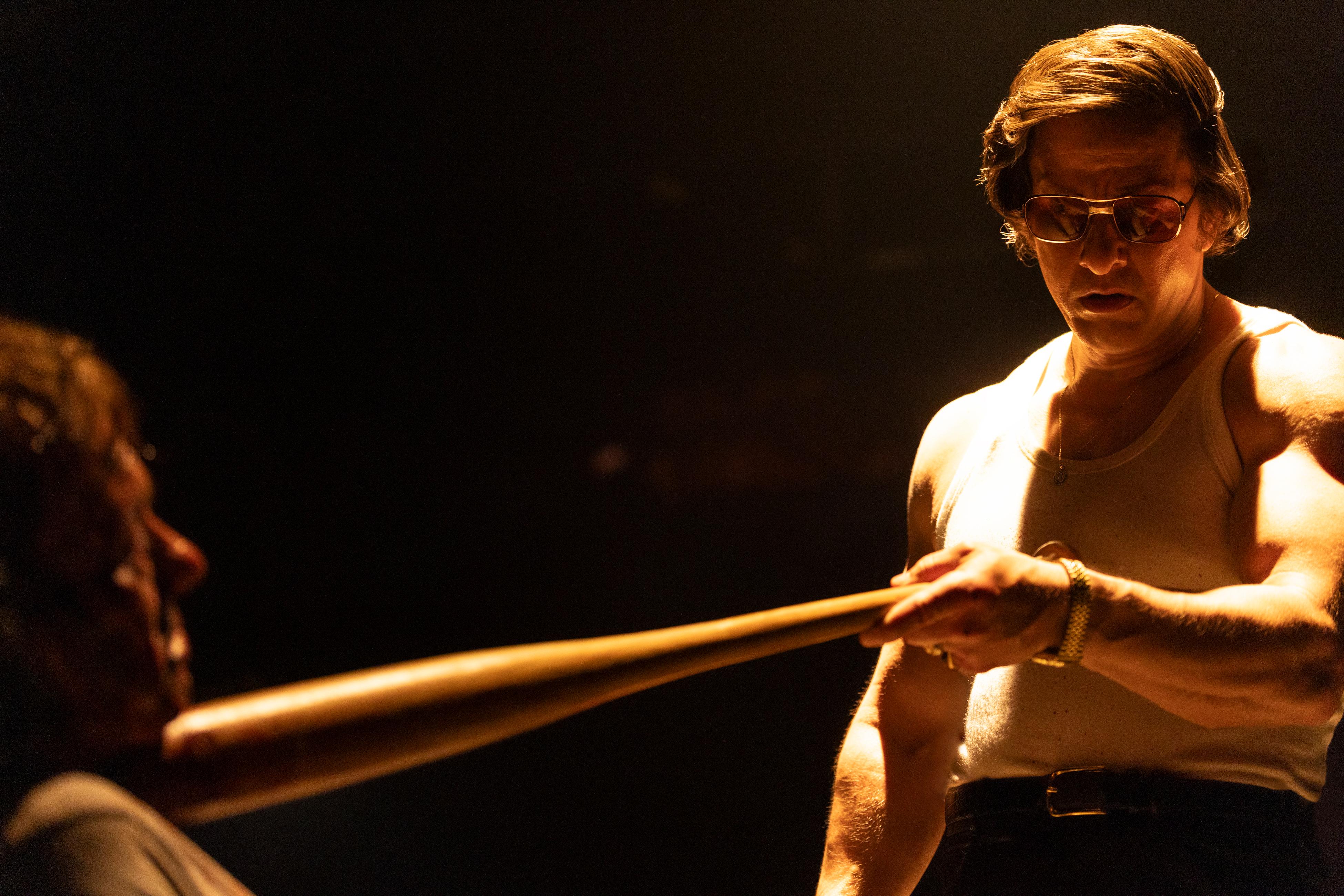 Mark Wahlberg, wearing sunglasses and a white undershirt, holds a wooden baseball bat toward another man.