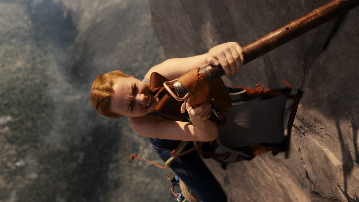 A red-haired climber grips a metal anchor while pressed against a sheer rock face.