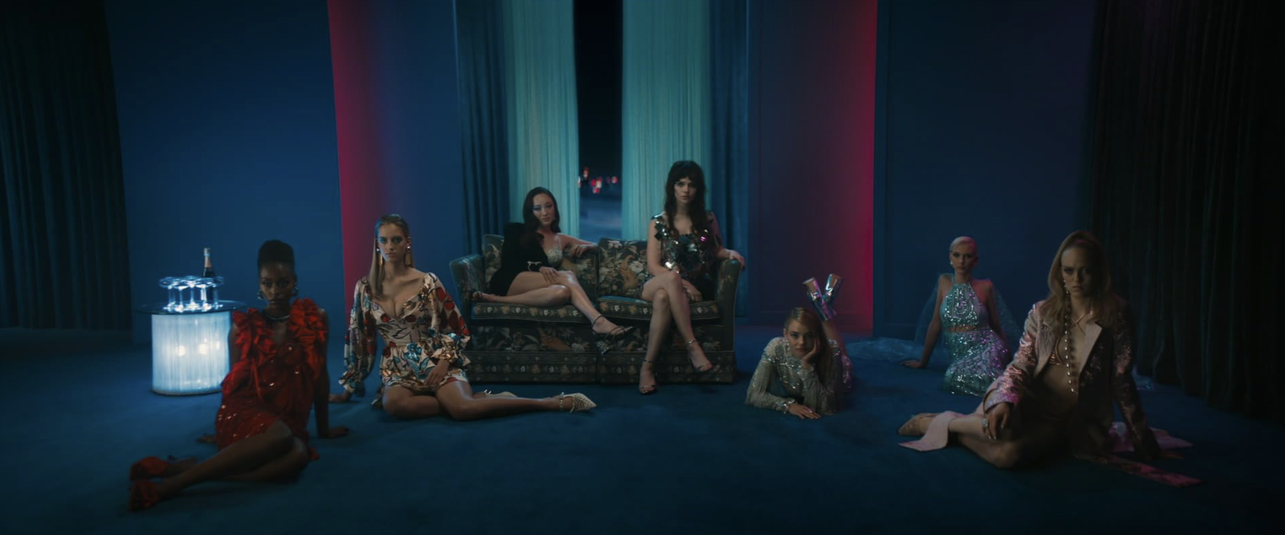 A group of women pose across a dark blue room illuminated by red and teal light.