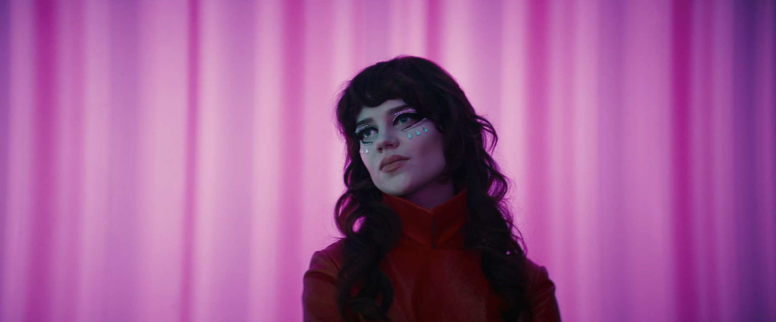 Sophie Thatcher wears a red high-collared outfit and dramatic eye makeup against a glowing pink background.