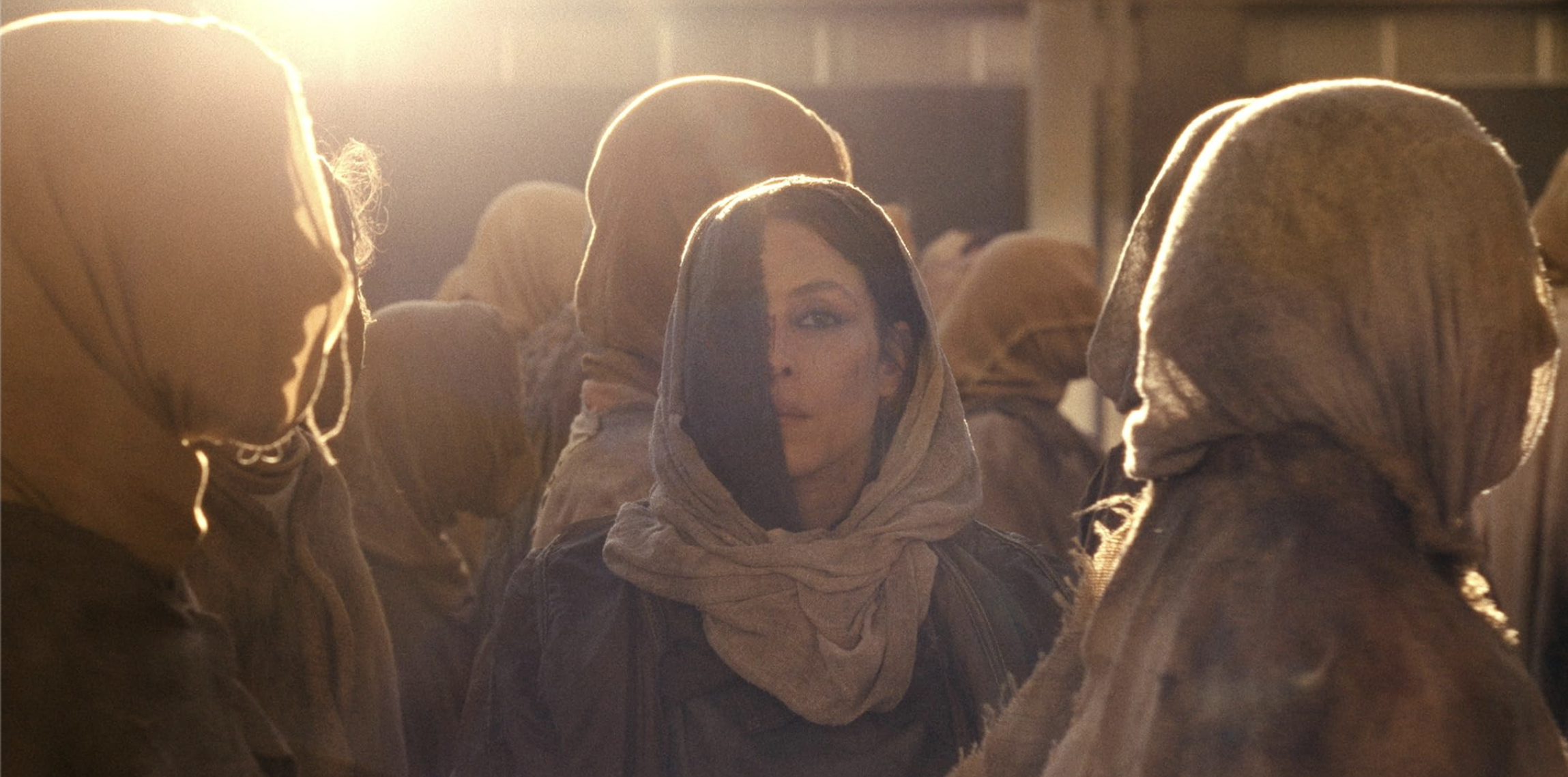 Noomi Rapace stands among hooded figures inside a warm, crowded space in Hot Spot.