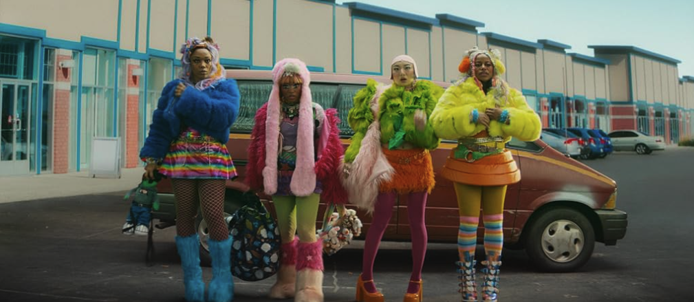 Four women in bright coordinated outfits stand in a parking lot beside a car.