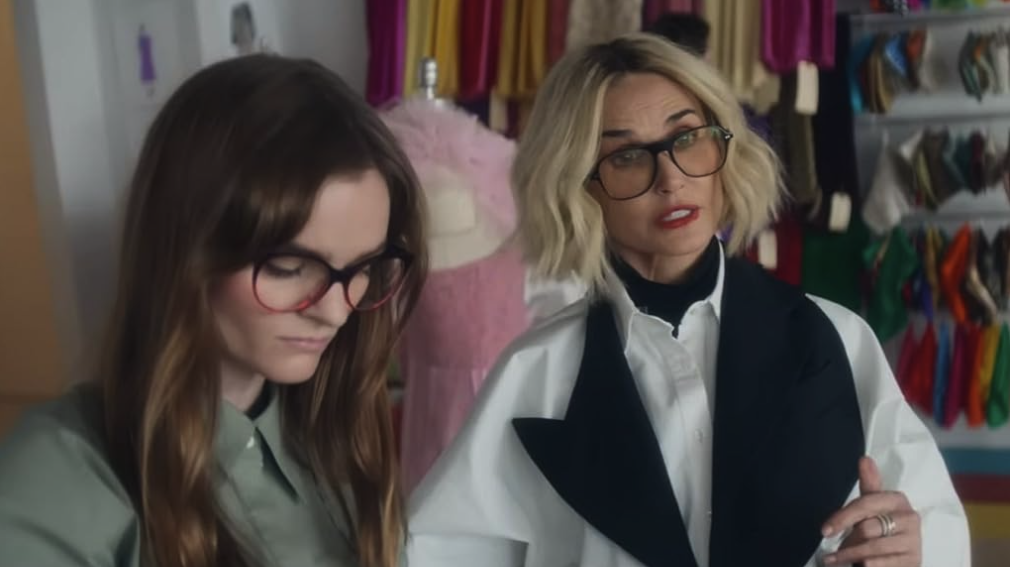Two women talk in a clothing store surrounded by racks of garments.