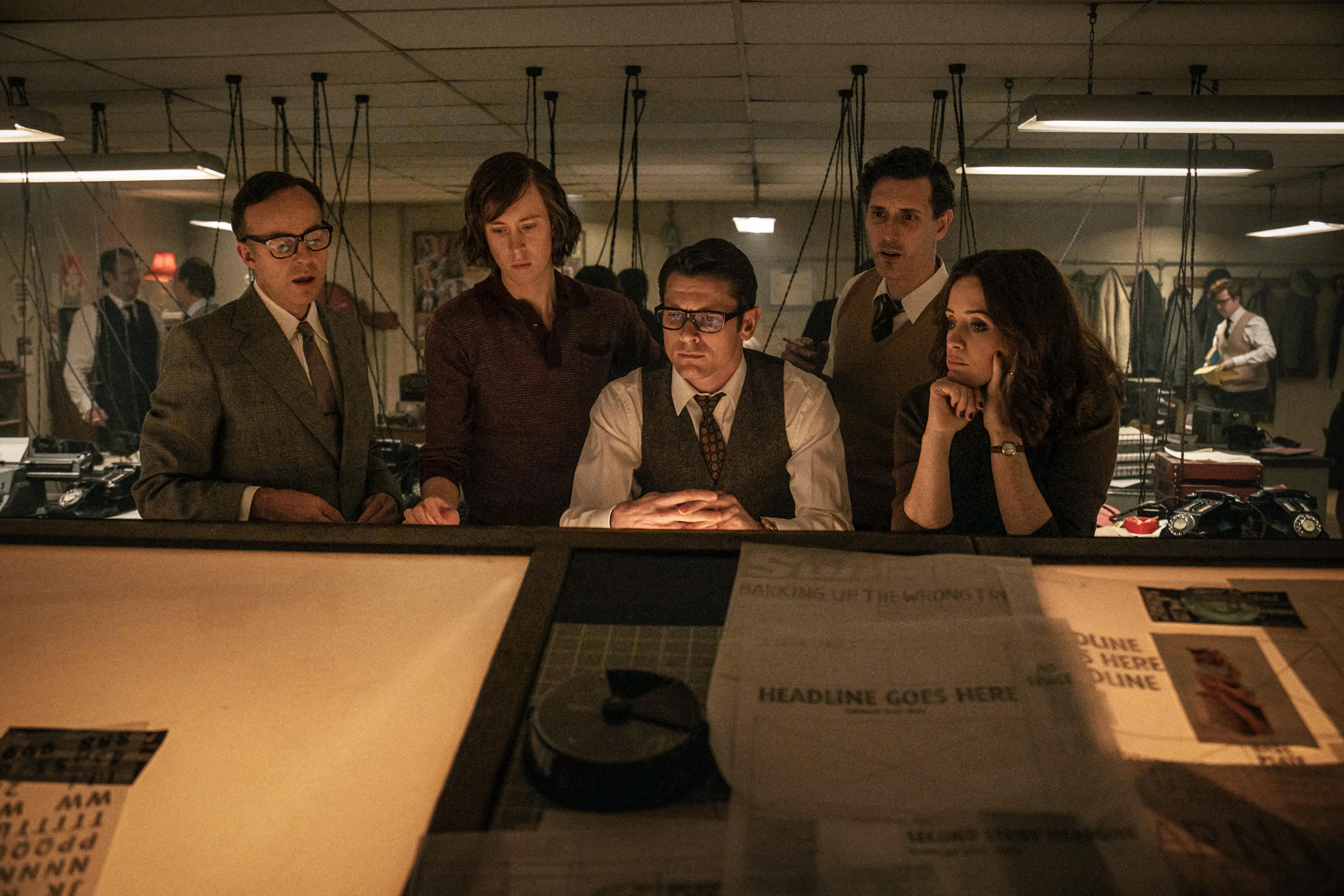 Newspaper staff gather around a desk in a crowded 1970s newsroom.