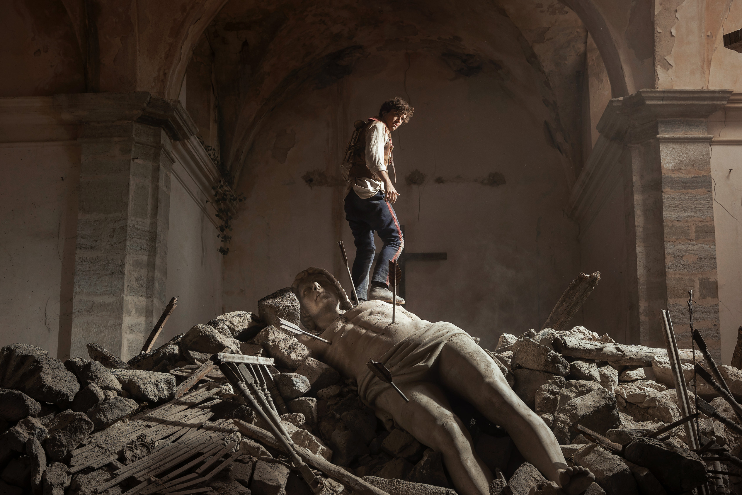 A young man stands on the remains of a fallen statue inside a ruined building