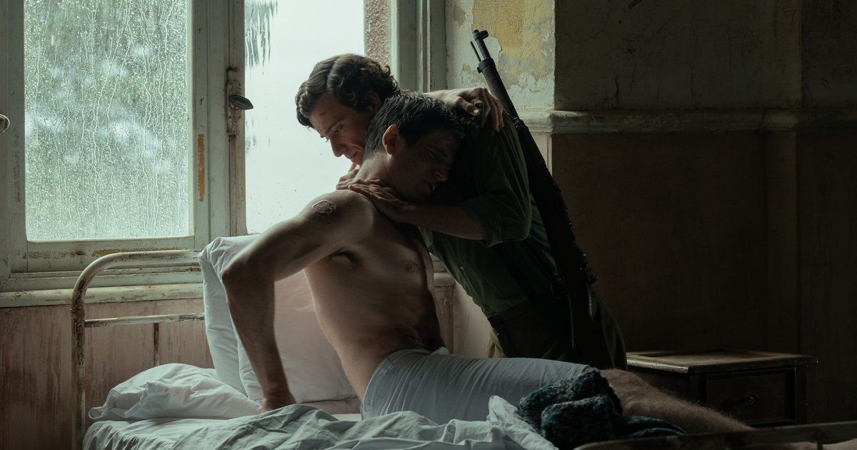 One man leans over another injured man sitting on a bed in a dim room.