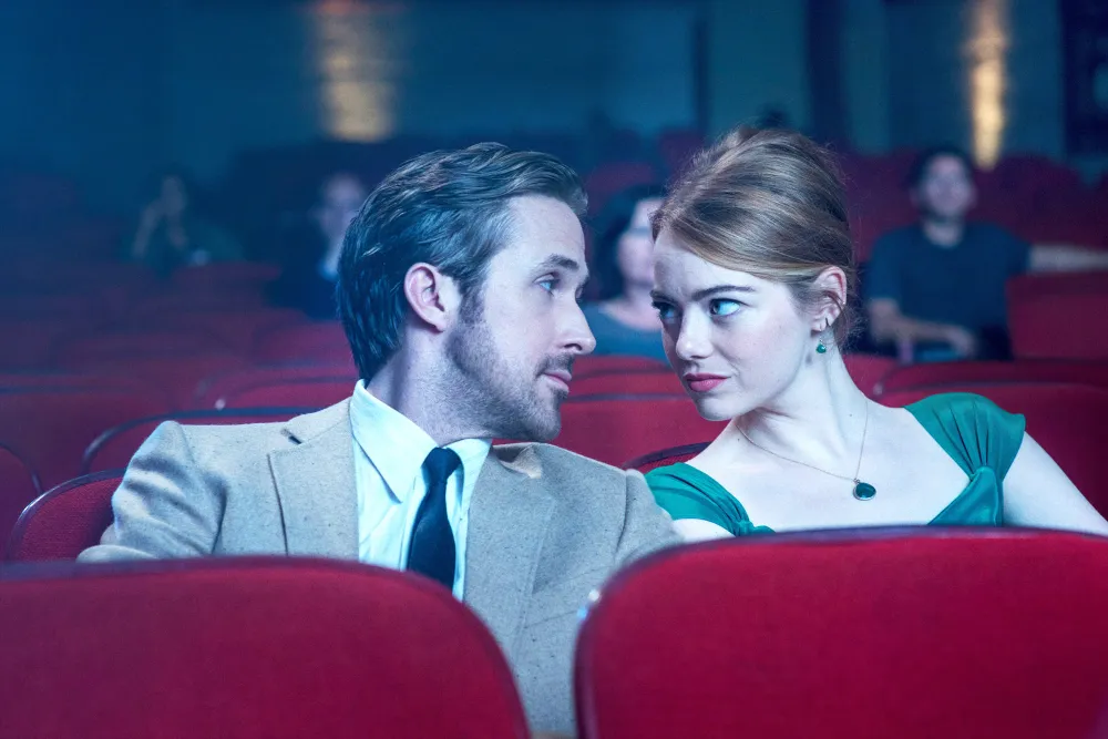 Ryan Gosling and Emma Stone sit close together in red theater seats in La La Land.