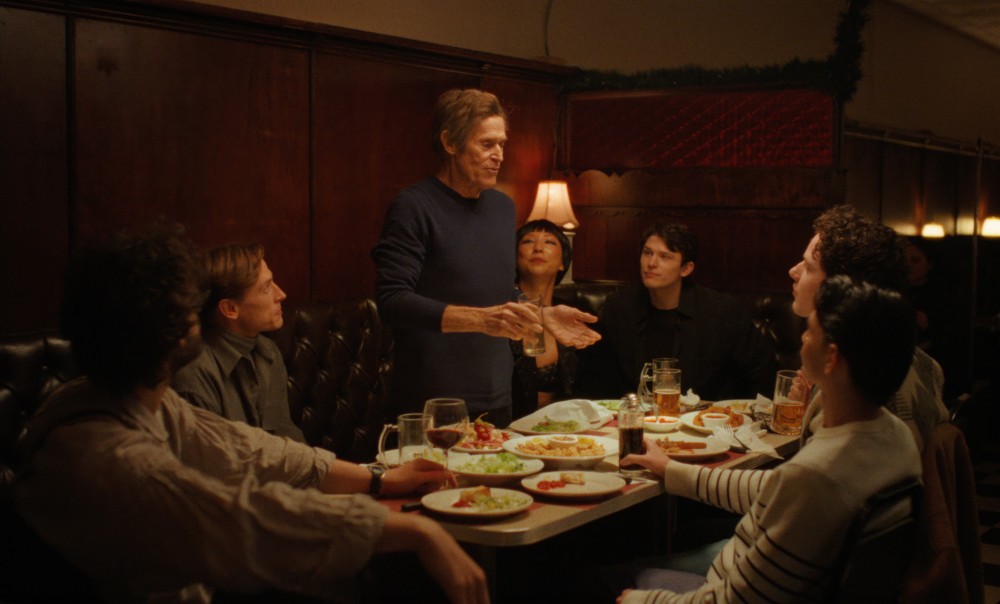 An older man stands speaking at a restaurant table while a group of younger diners watch him.