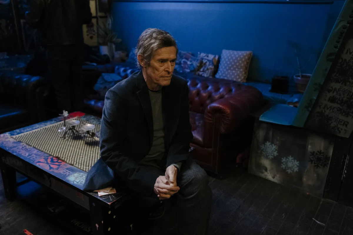 An older man sits alone in a dim blue room with his hands clasped in front of him.