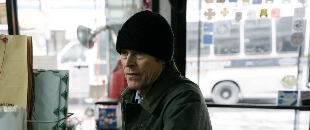 An older man wearing a black knit hat stands inside a small neighborhood store with a city bus visible through the window.