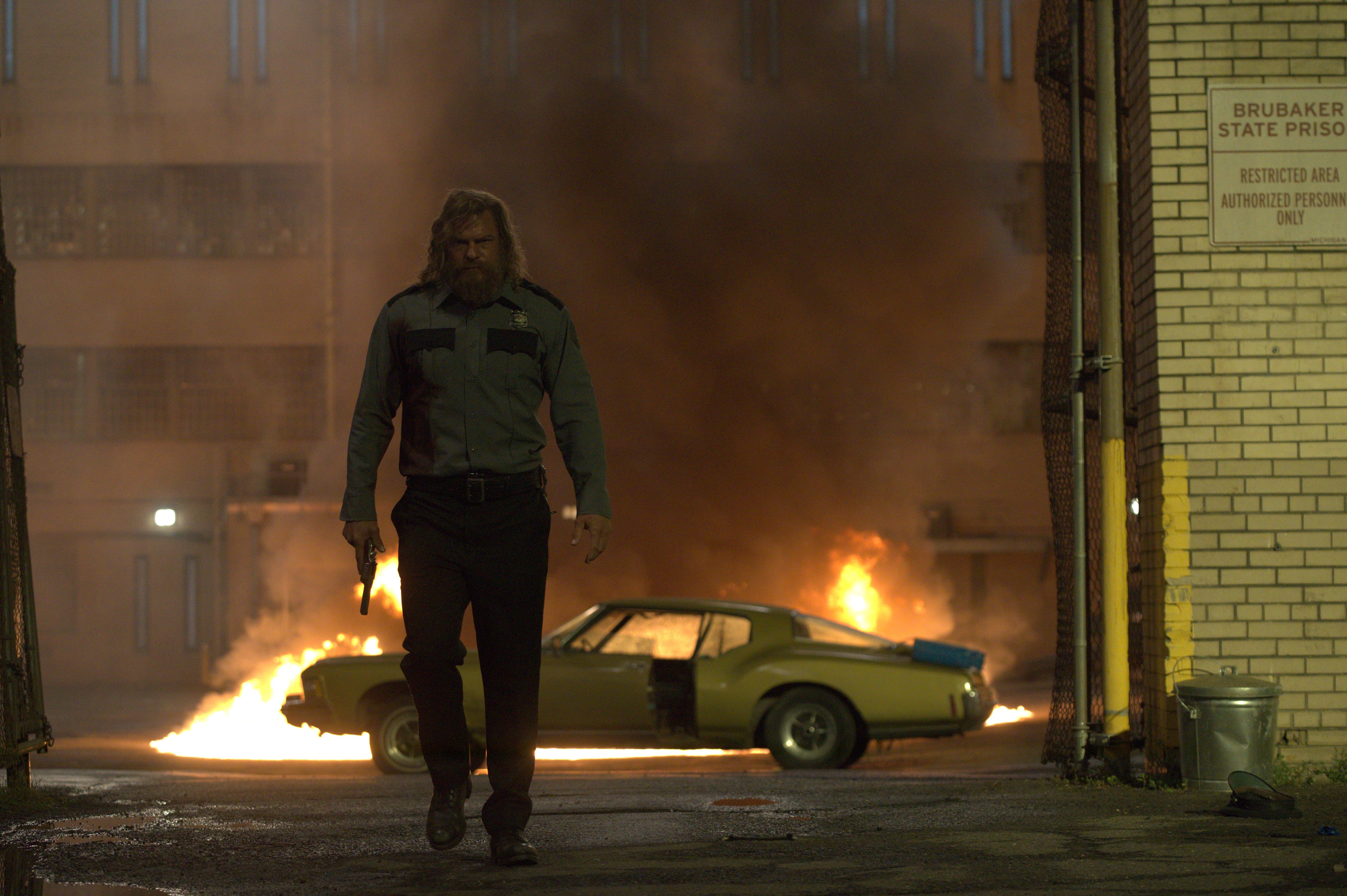 A bearded man in a prison guard uniform walks through an industrial yard while a car burns behind him.