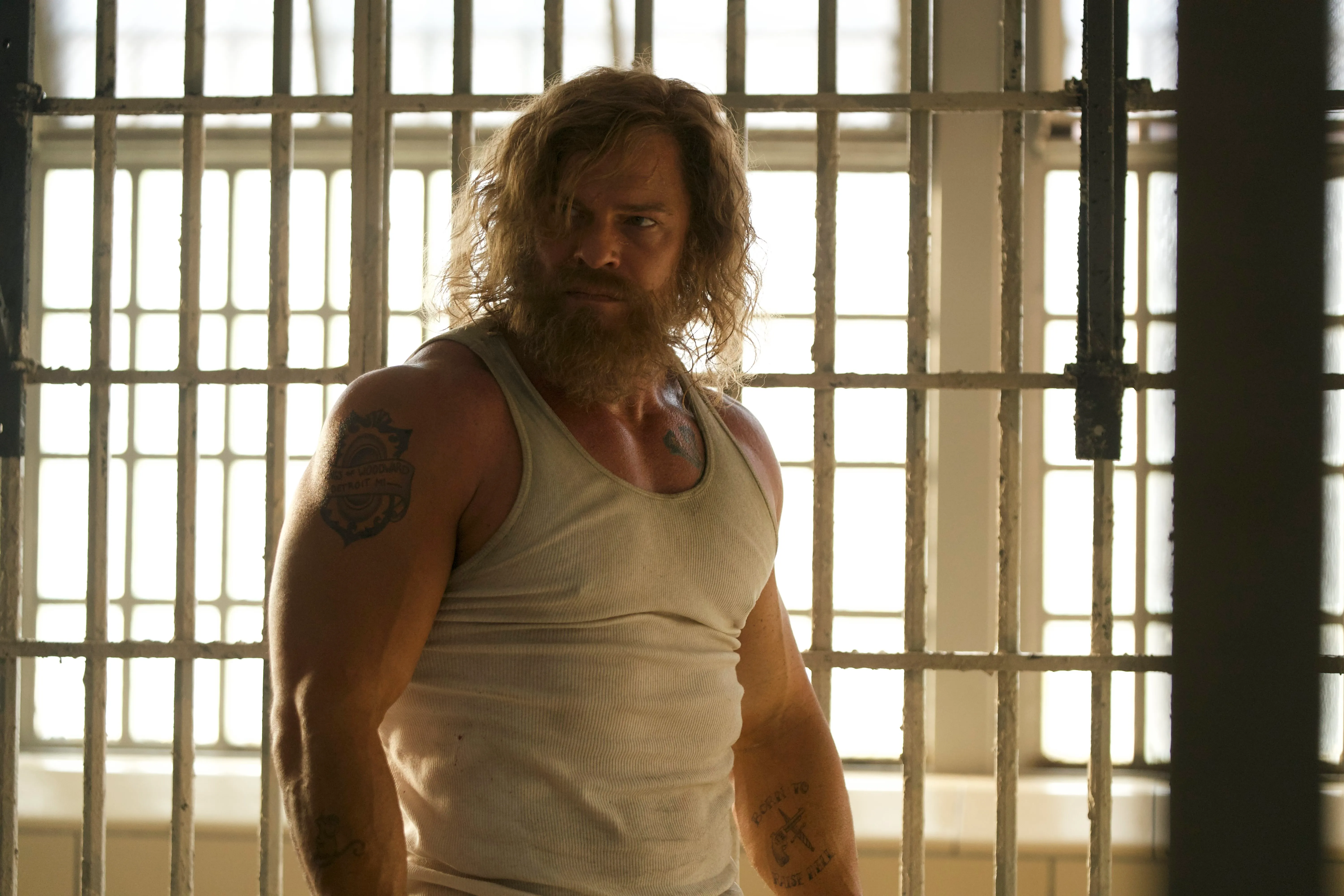 A muscular bearded man in a white undershirt stands inside a prison cell, framed by metal bars and bright windows.