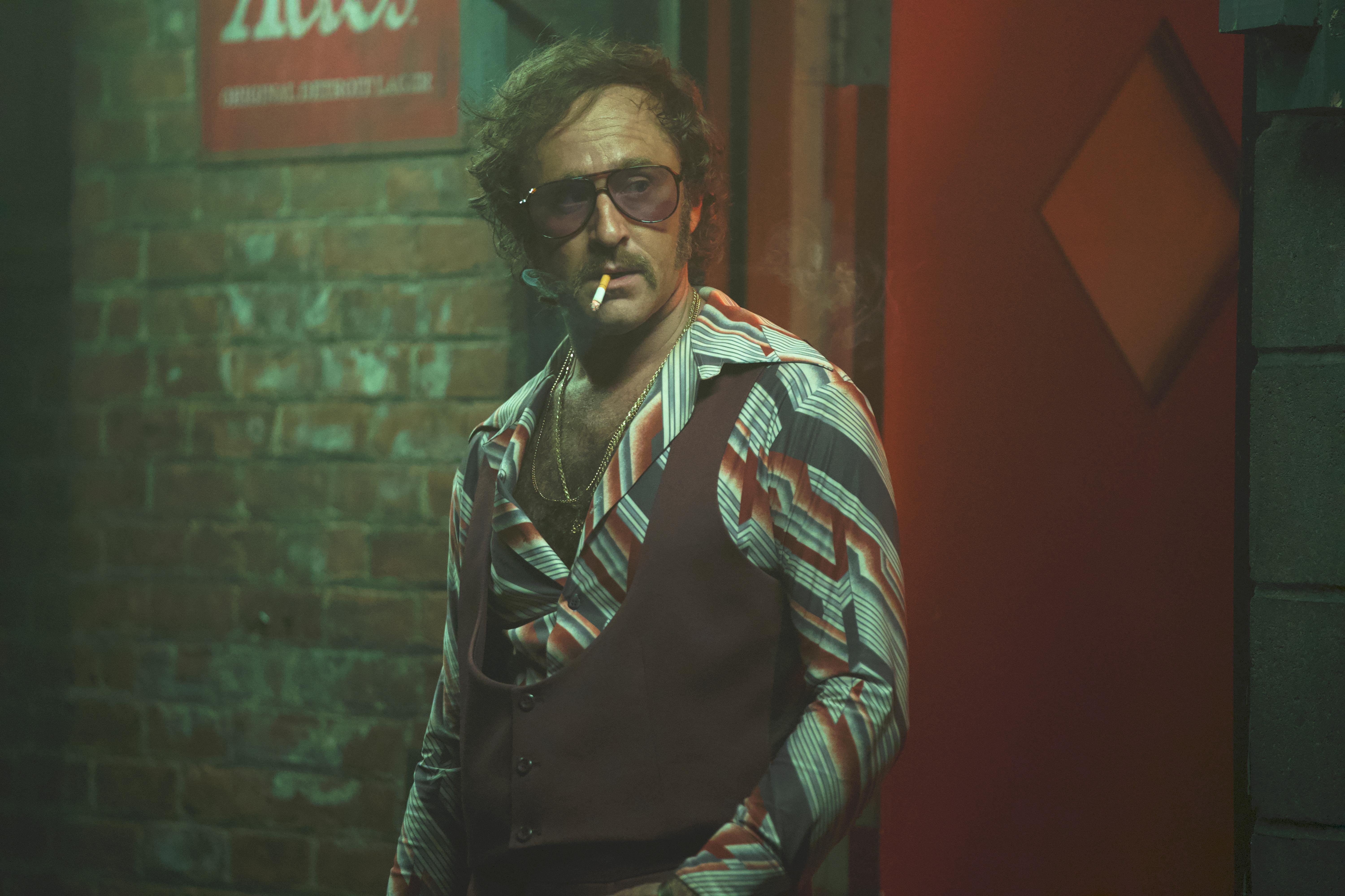 A mustached man wearing tinted glasses, a patterned shirt, and a dark vest stands in the doorway of a dim nightclub.