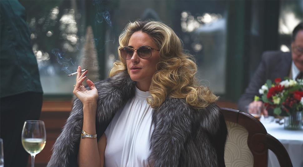 A blonde woman wearing sunglasses and a fur coat sits at a restaurant table while holding a cigarette.
