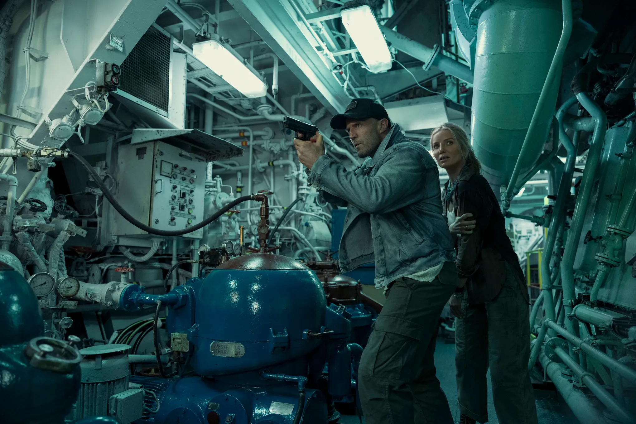 Jason Statham aims a handgun while Annabelle Wallis stands behind him inside the machinery of a cargo ship in Mutiny.