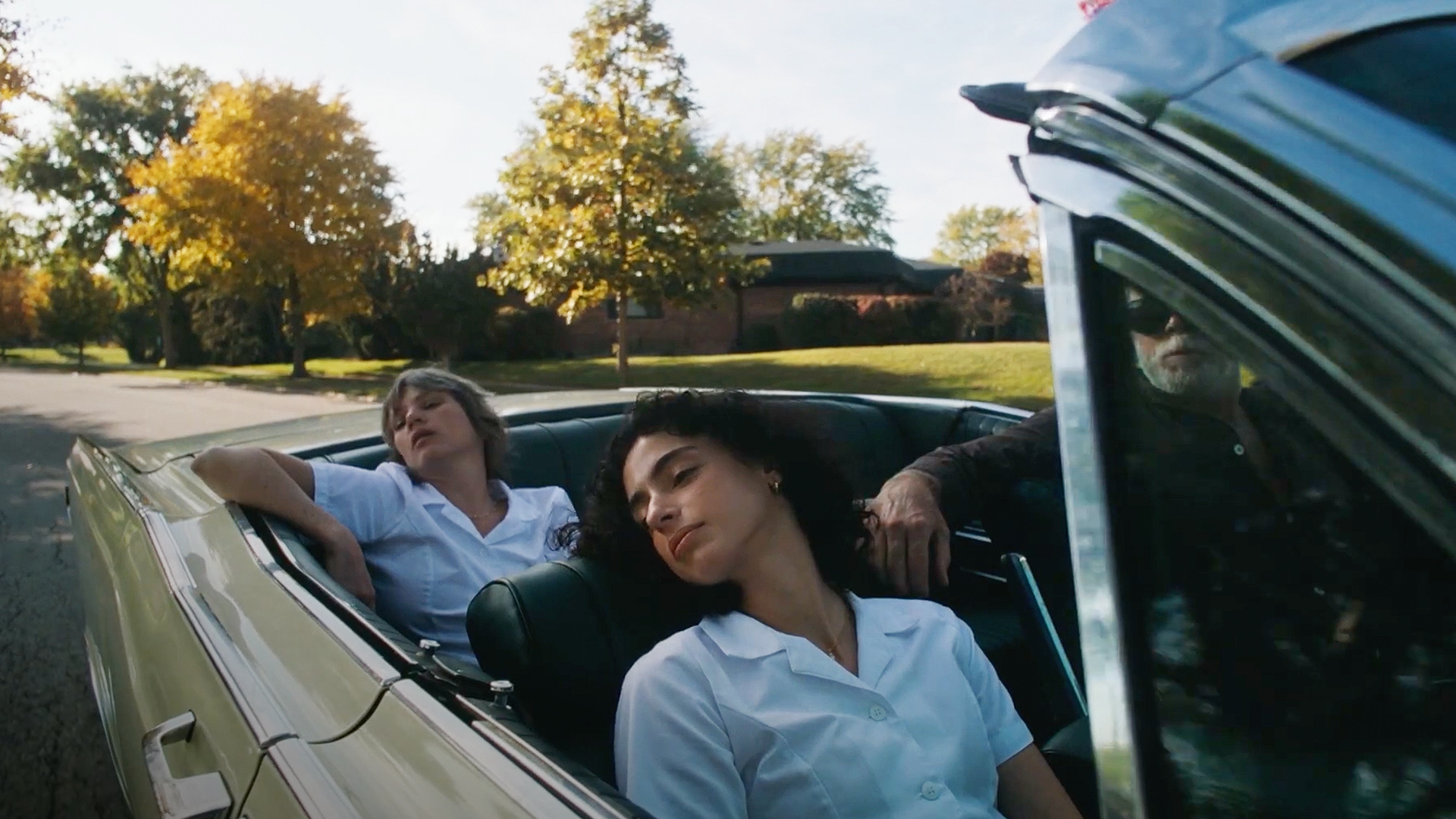 Two women in white nursing uniforms recline in the back seat of a convertible on a quiet suburban street.