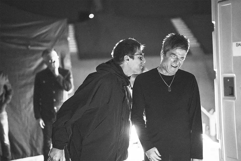 Liam Gallagher leans toward a smiling Noel Gallagher backstage in a black-and-white photograph.