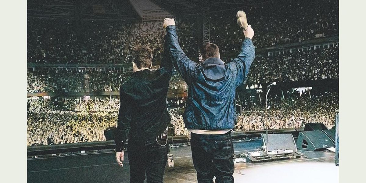 Noel and Liam Gallagher stand with their backs to the camera and raise their joined hands before a packed stadium crowd.
