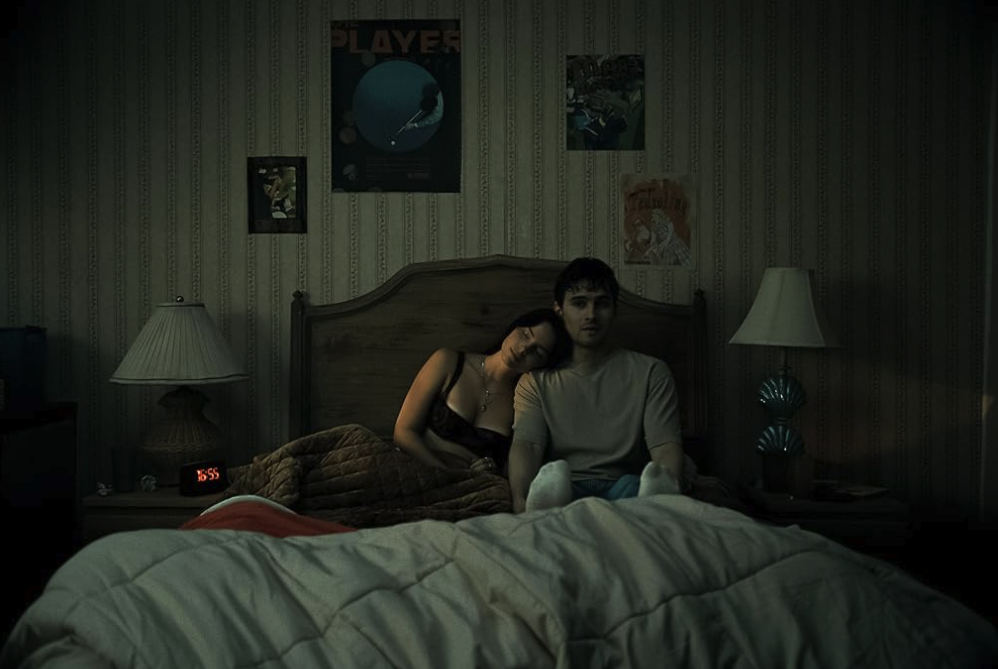 A couple sit together on a bed in a dimly lit bedroom covered in posters.