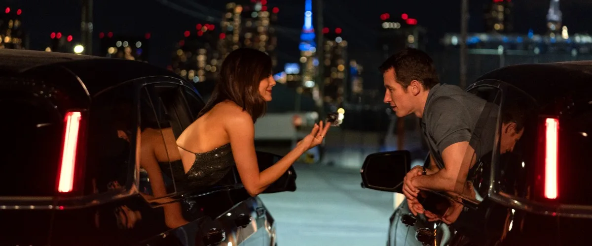 Monica Barbaro and Callum Turner lean toward each other from separate cars at night with the New York skyline behind them.