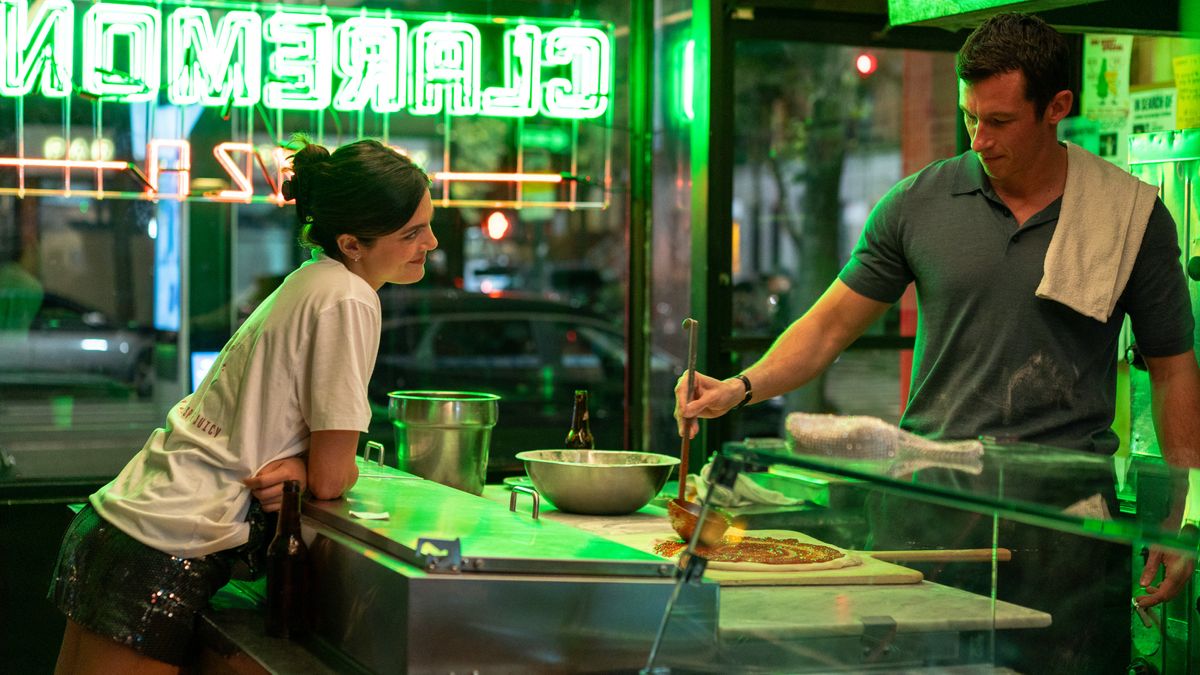 Monica Barbaro leans across the counter of a neon-lit pizza shop while Callum Turner prepares a pizza behind the counter.